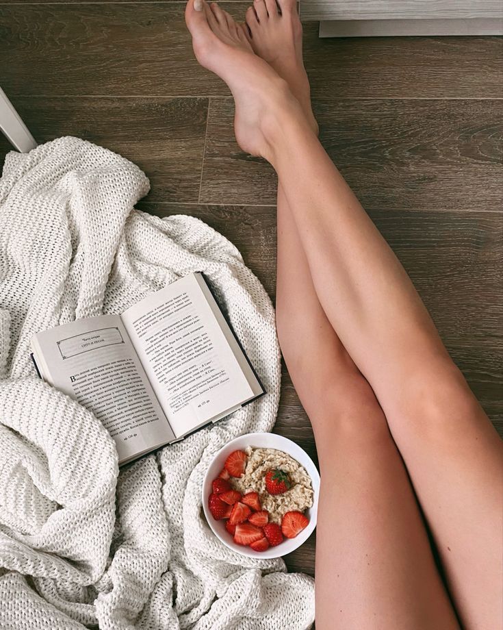Bare legs on a wooden floor with a book, blanket, and bowl of oatmeal with strawberries