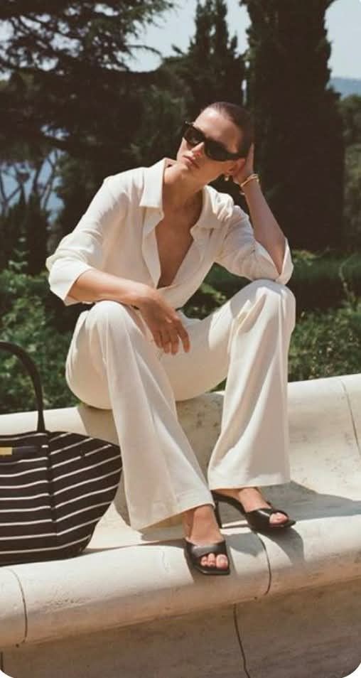 Woman in a white pantsuit and sunglasses sitting outdoors with a striped bag