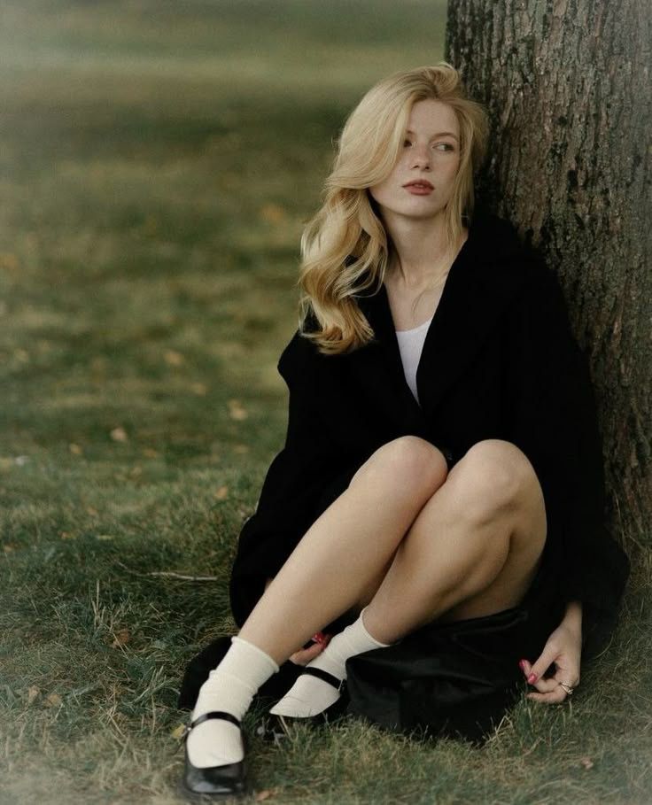 Blonde woman in a black coat and white socks sitting on grass next to a tree