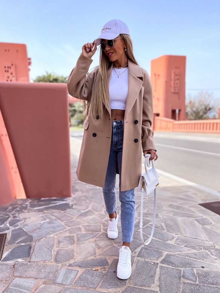 Woman in a beige coat, crop top, and jeans walking outdoors