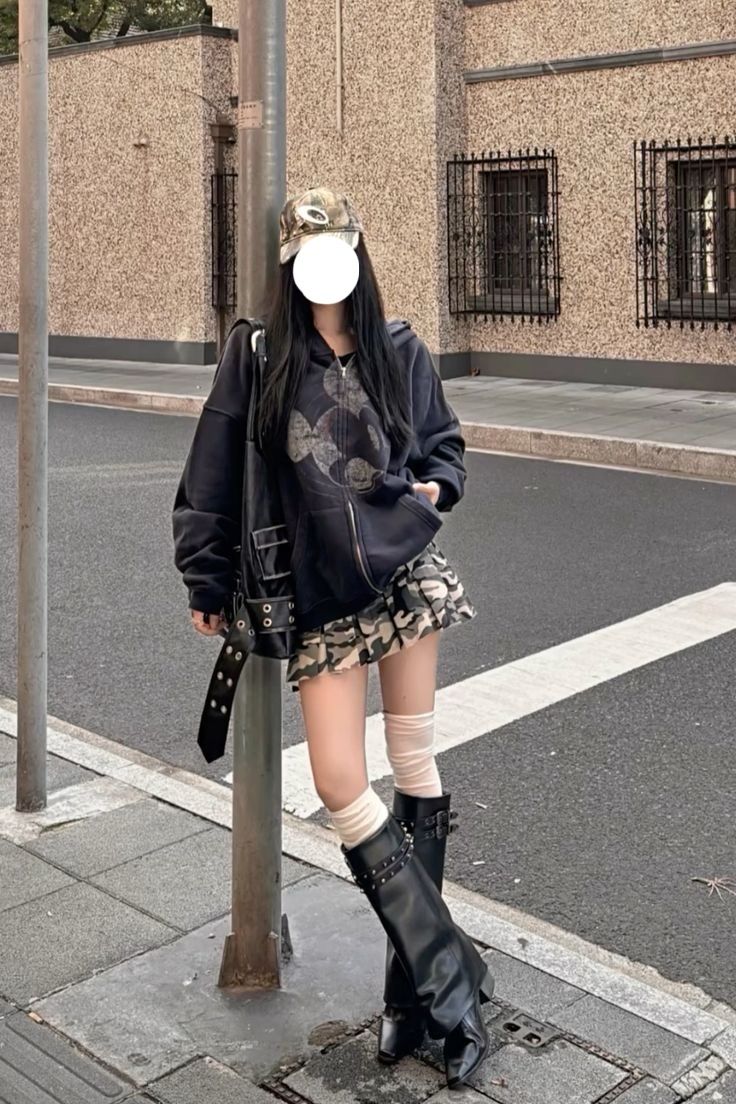 Person in a black hoodie and camo skirt leaning against a pole on a street