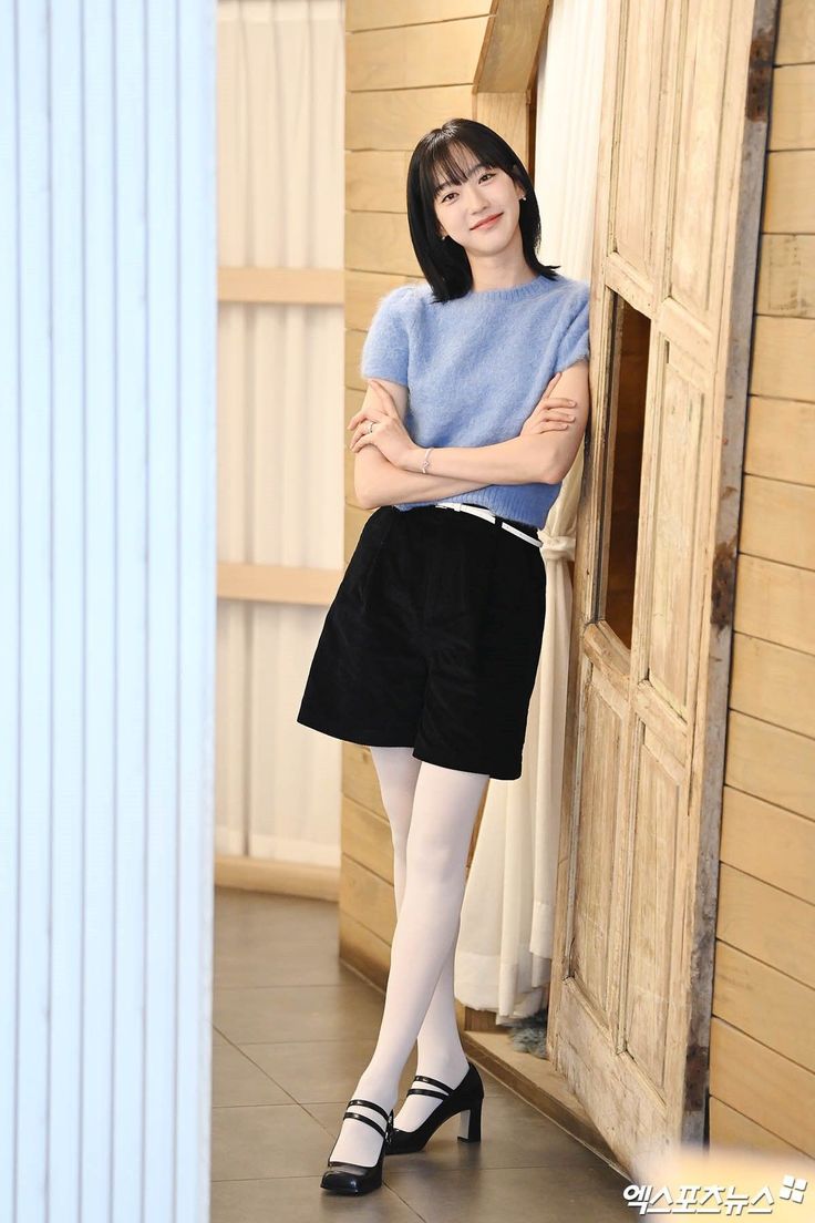 Woman with short black hair wearing a blue sweater and black shorts with white tights