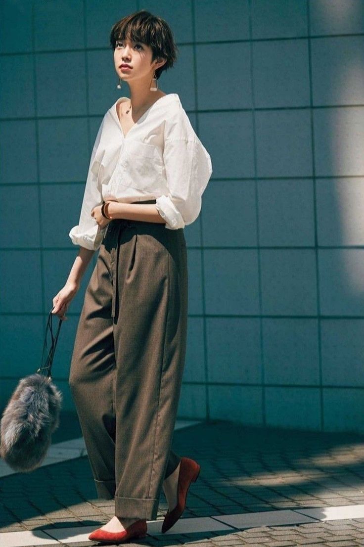 Woman with short hair in a white shirt and wide leg pants holding a furry bag