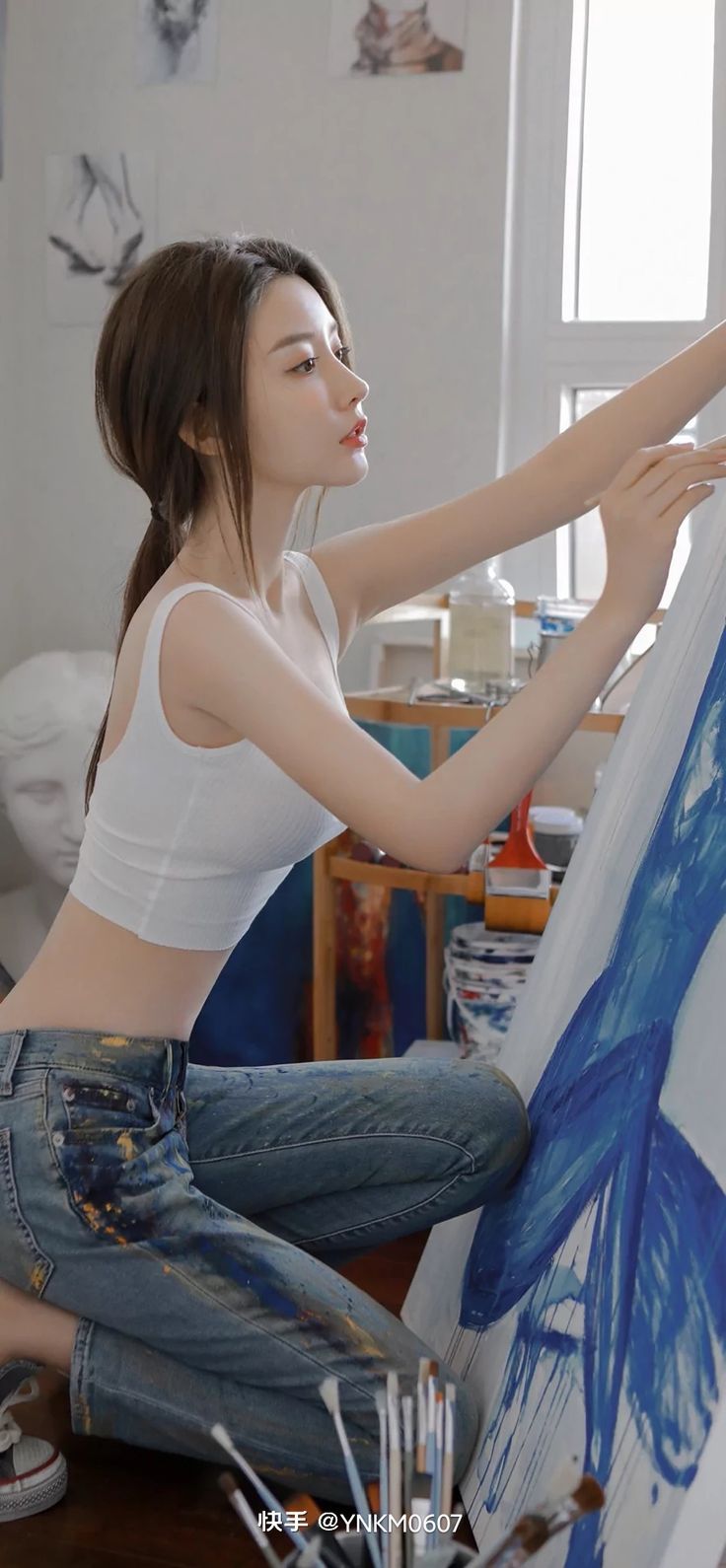 Woman in a white crop top and paint-splattered jeans painting in an art studio