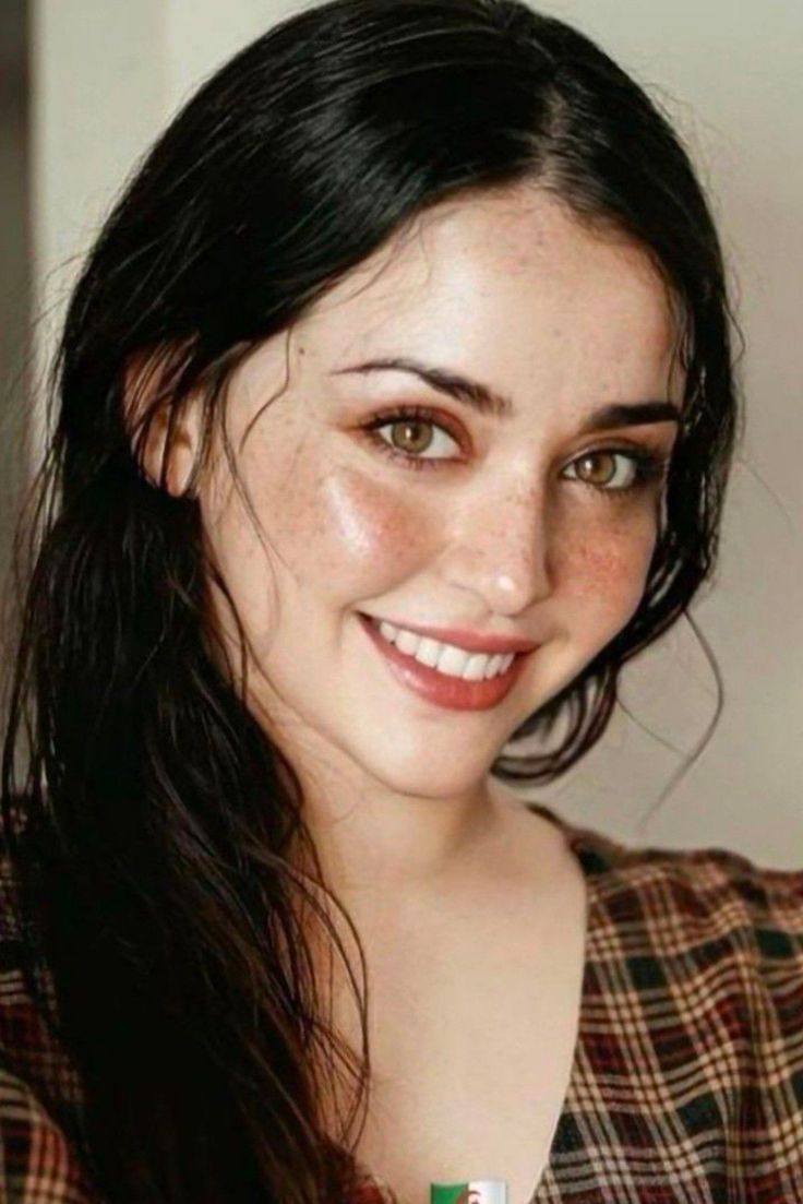 Woman with long dark hair and freckles smiling in a plaid shirt