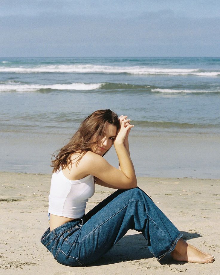 Woman with long hair sitting on a sandy beach in a white tank top and blue jeans