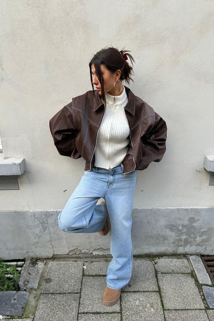 Woman in a brown leather jacket and light wash jeans leaning against a wall
