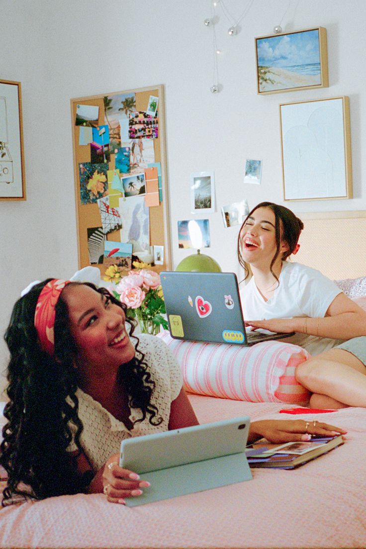 Two young women laughing and relaxing on a bed with a laptop and tablet