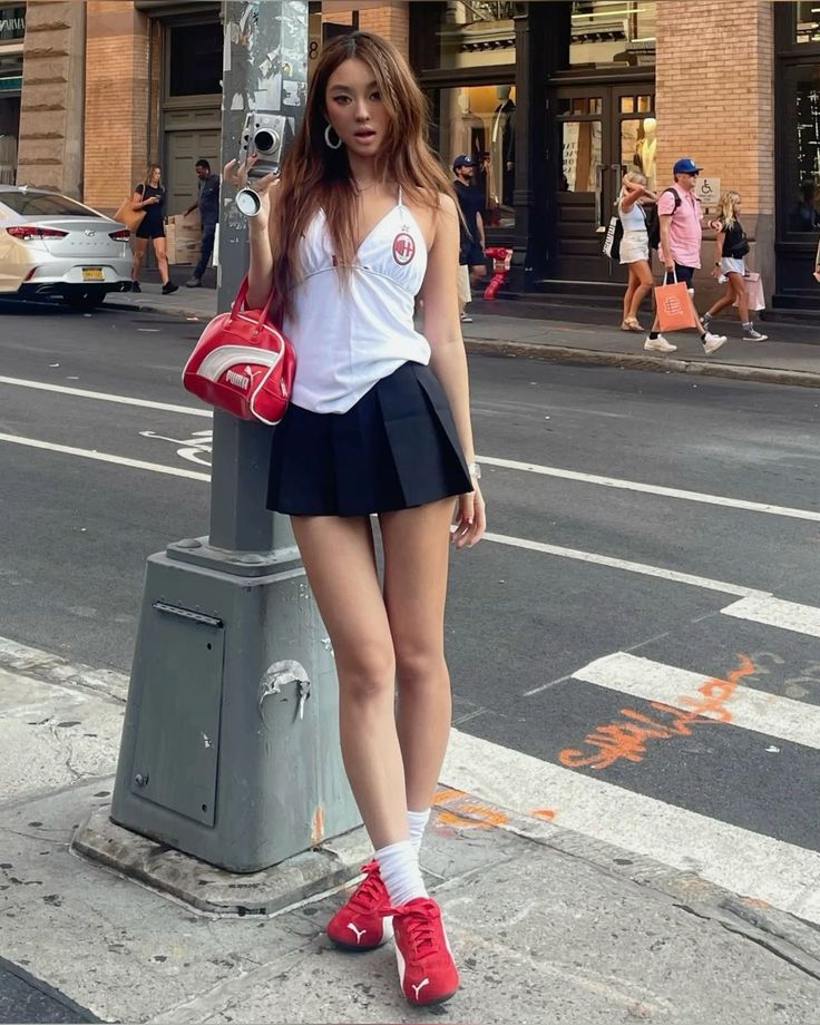 Woman in a white top and black pleated skirt holding a camera on a city street