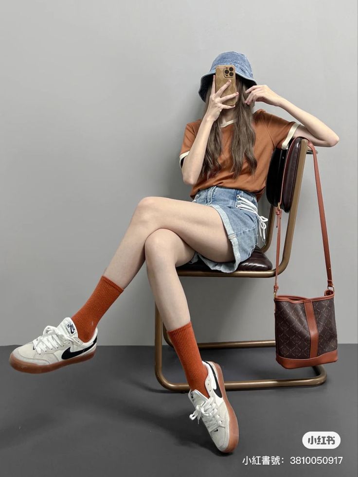 Person in a denim bucket hat and shorts sitting on a chair, taking a mirror selfie