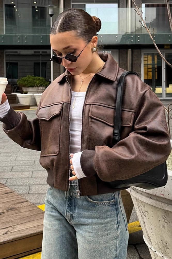 Woman in a brown leather jacket and sunglasses holding a cup outdoors