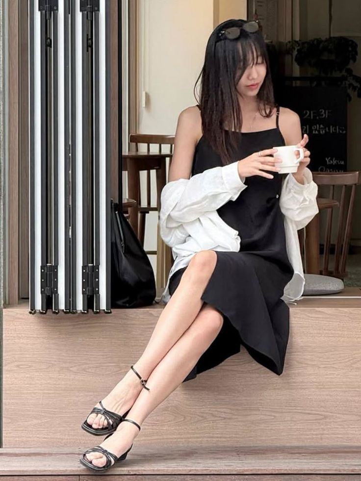 Woman in a black dress and white shirt sitting outdoors, holding a cup