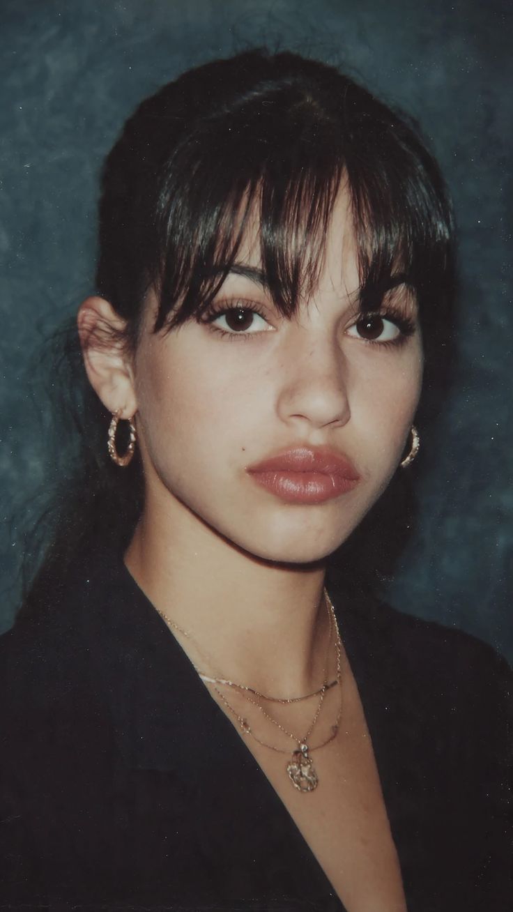 Woman with bangs and hoop earrings wearing layered necklaces and a black top