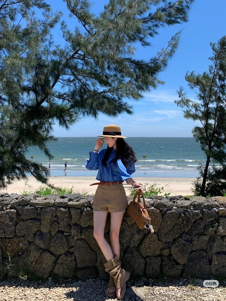 Woman in a straw hat and blue shirt standing by a stone wall at the beach
