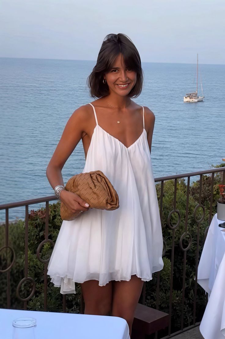 Woman in a white dress holding a brown bag with an ocean view and boat in the background