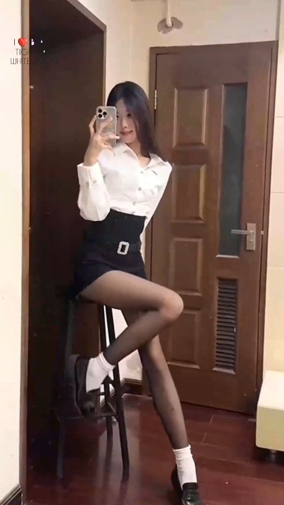Woman in a white shirt and black skirt taking a mirror selfie indoors