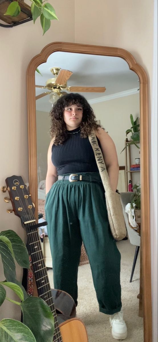 Woman with curly hair in a black top and green pants taking a mirror selfie
