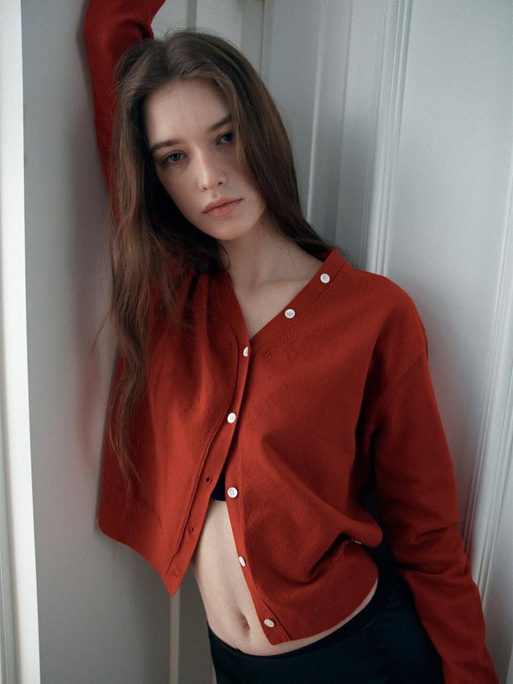 Woman in a red cardigan and black bra leaning against a white wall