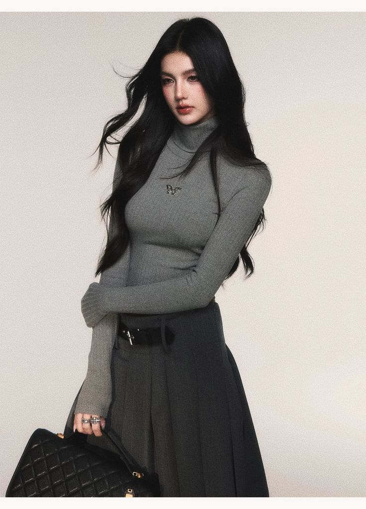 Woman with long black hair wearing a gray turtleneck and black skirt, holding a handbag