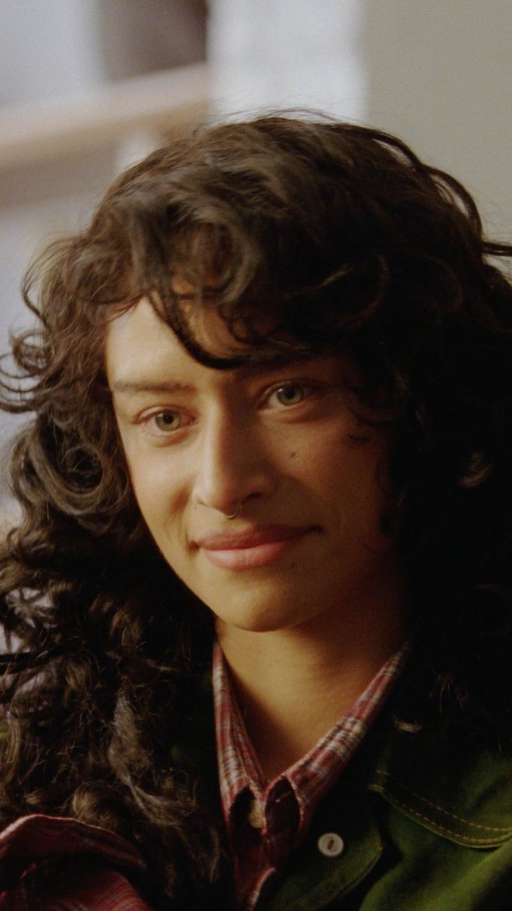 Person with curly hair and a nose ring wearing a plaid shirt and green jacket