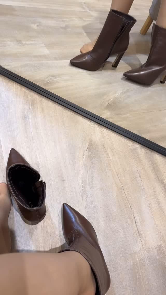 Person trying on brown high-heeled boots on a wooden floor with a mirror reflection