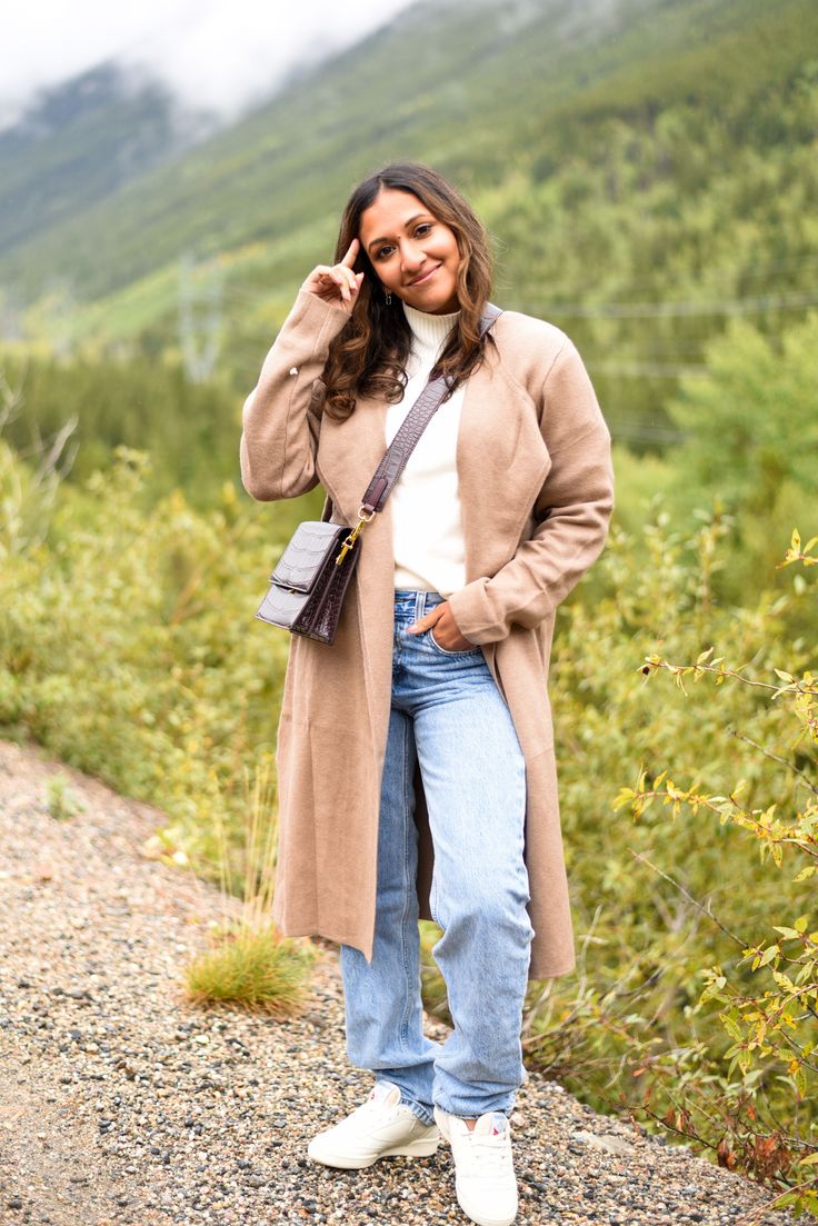Woman in a long coat and jeans posing outdoors with a mountain background