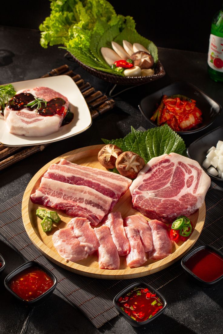 Assortment of raw pork cuts, kimchi, and vegetables on a dark table