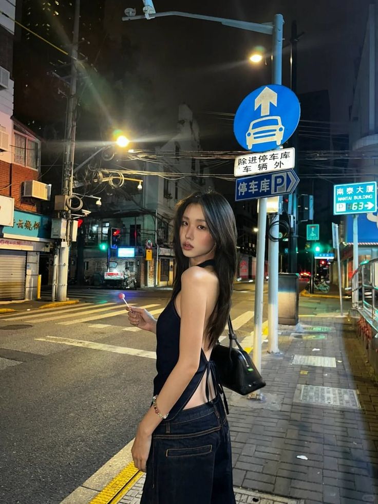 Woman in a backless top and jeans holding a lollipop on a city street at night