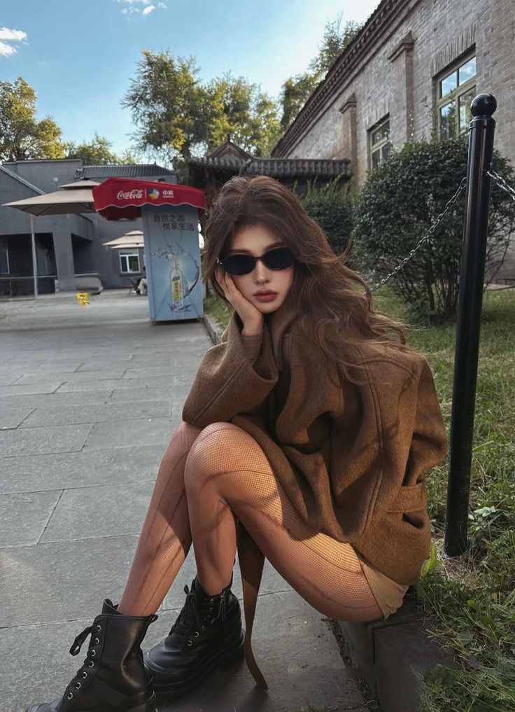 Woman with long hair and sunglasses sitting outdoors in a brown coat and fishnet stockings