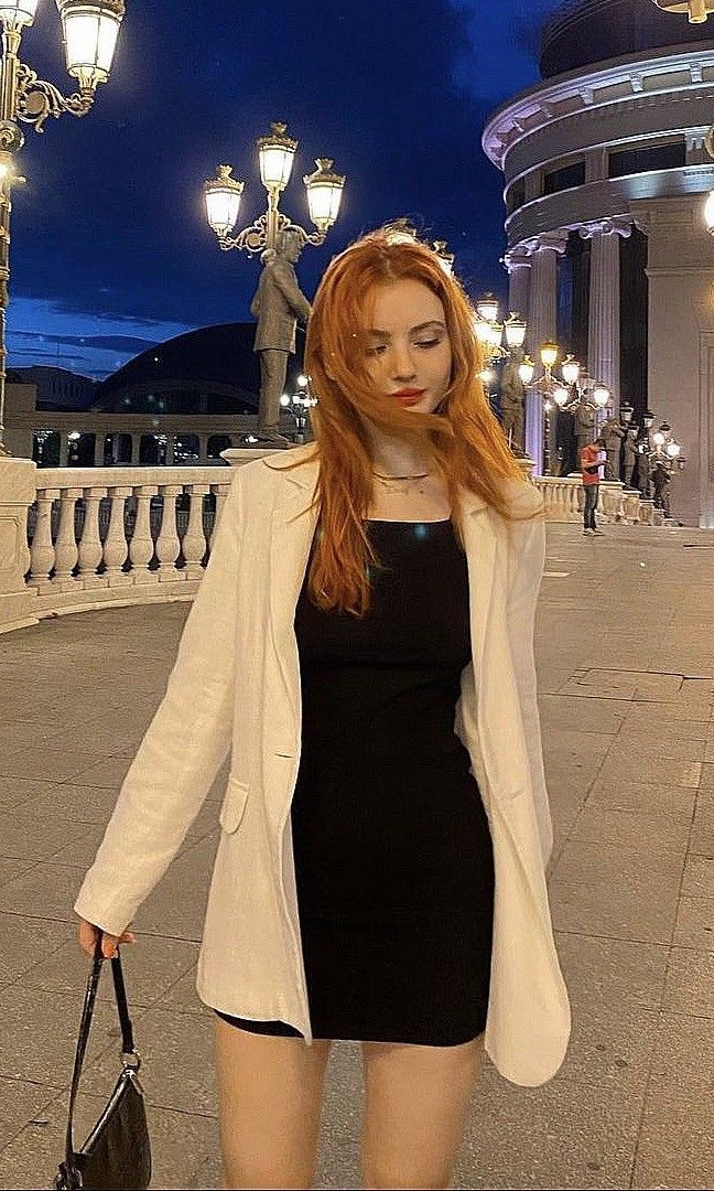 Woman with long red hair in a black dress and white blazer on a city street at night