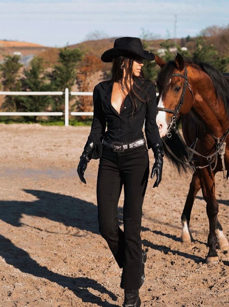 Woman in a cowboy hat and black outfit walking with a horse outdoors