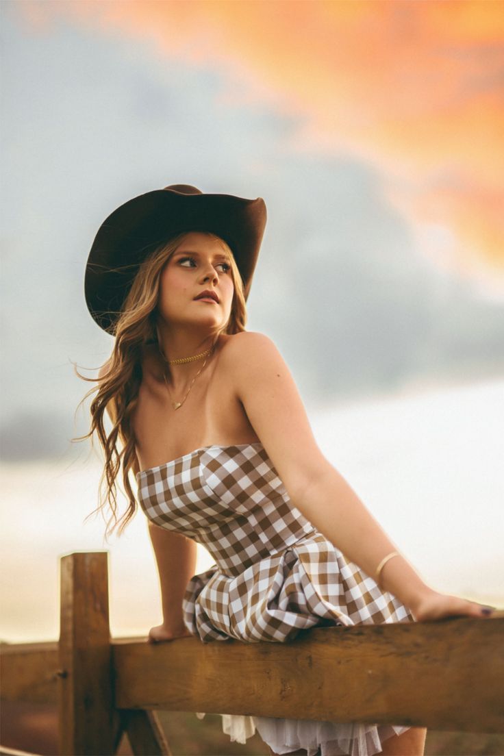 Woman in a cowboy hat and gingham dress sitting on a wooden fence at sunset