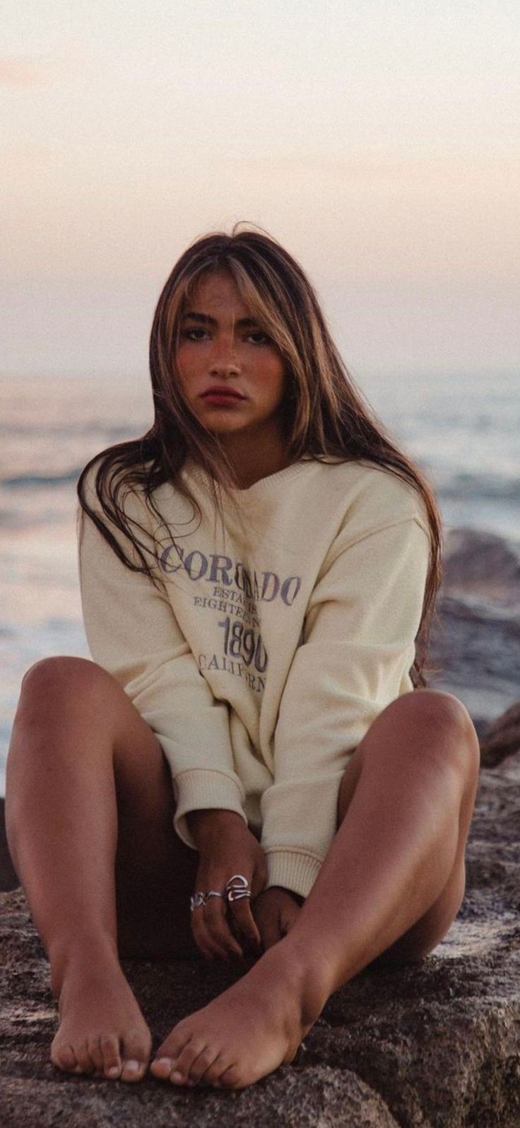 Woman with long hair wearing a sweatshirt sitting on rocks at sunset