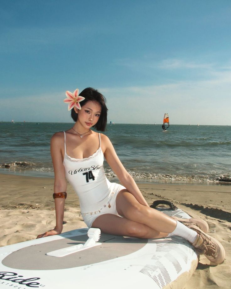 Woman in a white swimsuit with a flower in her hair sitting on a surfboard at the beach