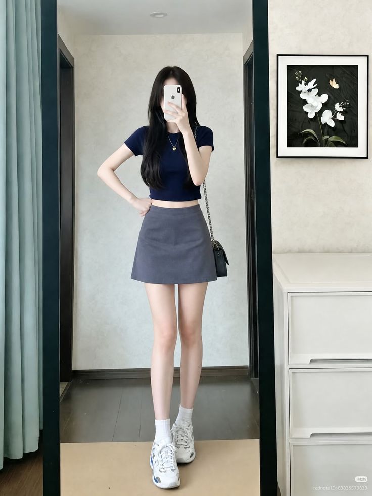 Woman in a crop top and mini skirt taking a mirror selfie indoors
