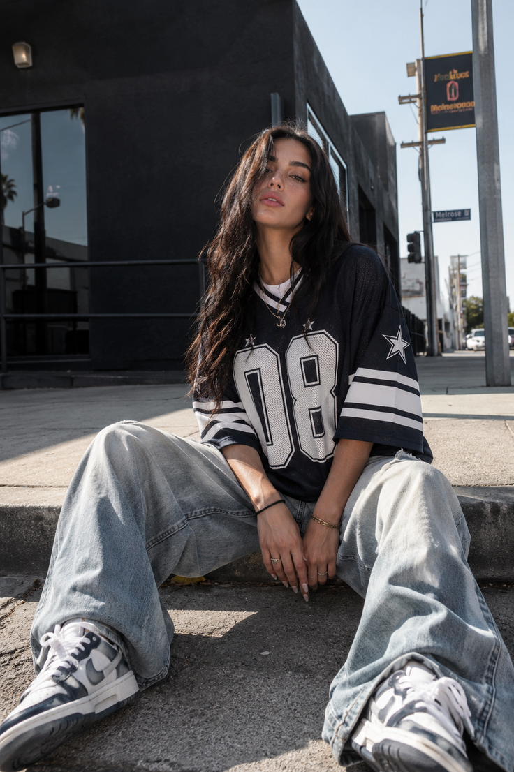 Woman with long dark hair wearing an oversized jersey and baggy jeans, sitting on a curb