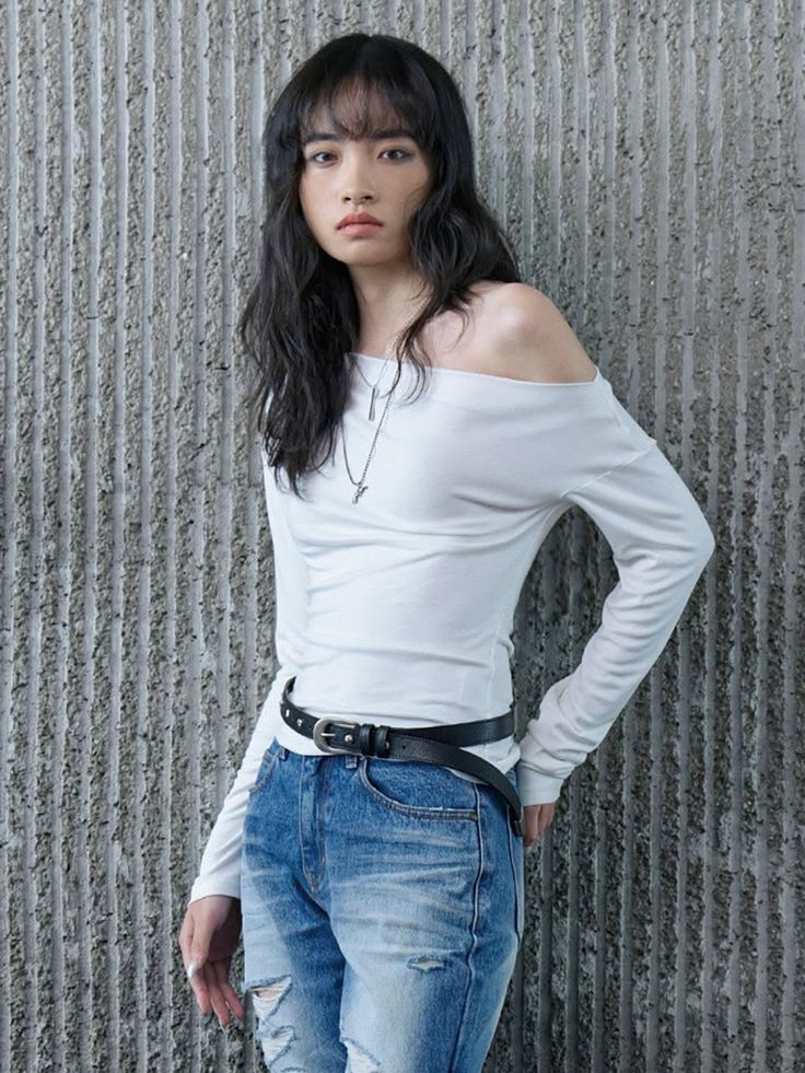 Person with long dark hair wearing an off-shoulder top and ripped jeans indoors