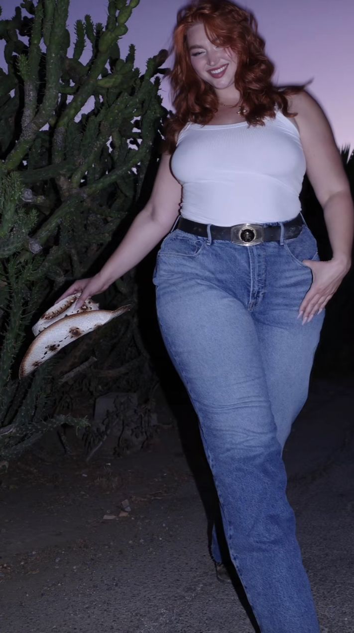 Woman with red hair in a white tank top and blue jeans holding a cowboy hat outdoors at night
