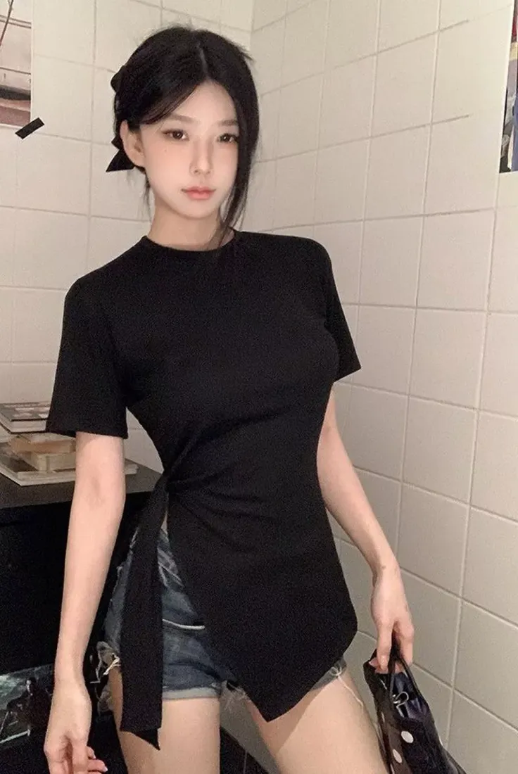 Woman with black hair wearing a black side-tie top and denim shorts indoors