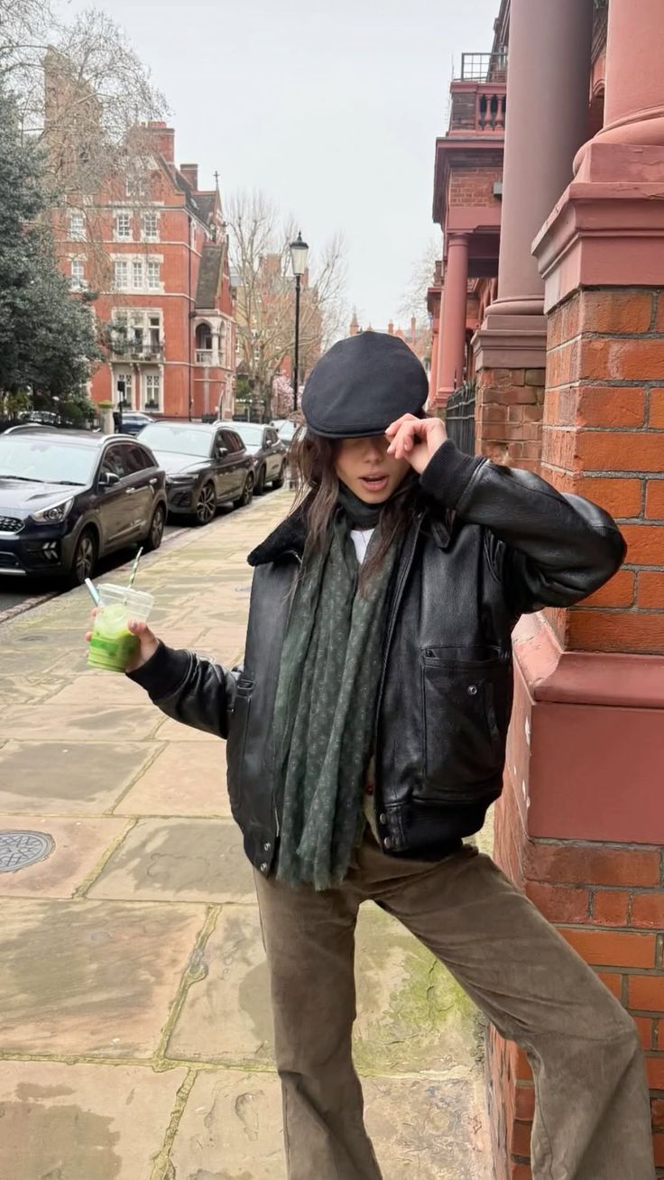 Woman in a black leather jacket and flat cap holding a green drink on a street