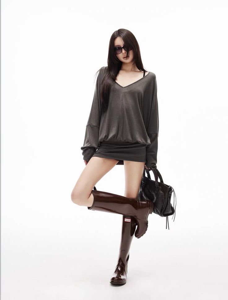 Woman in an oversized top, mini skirt, and rain boots posing with a handbag