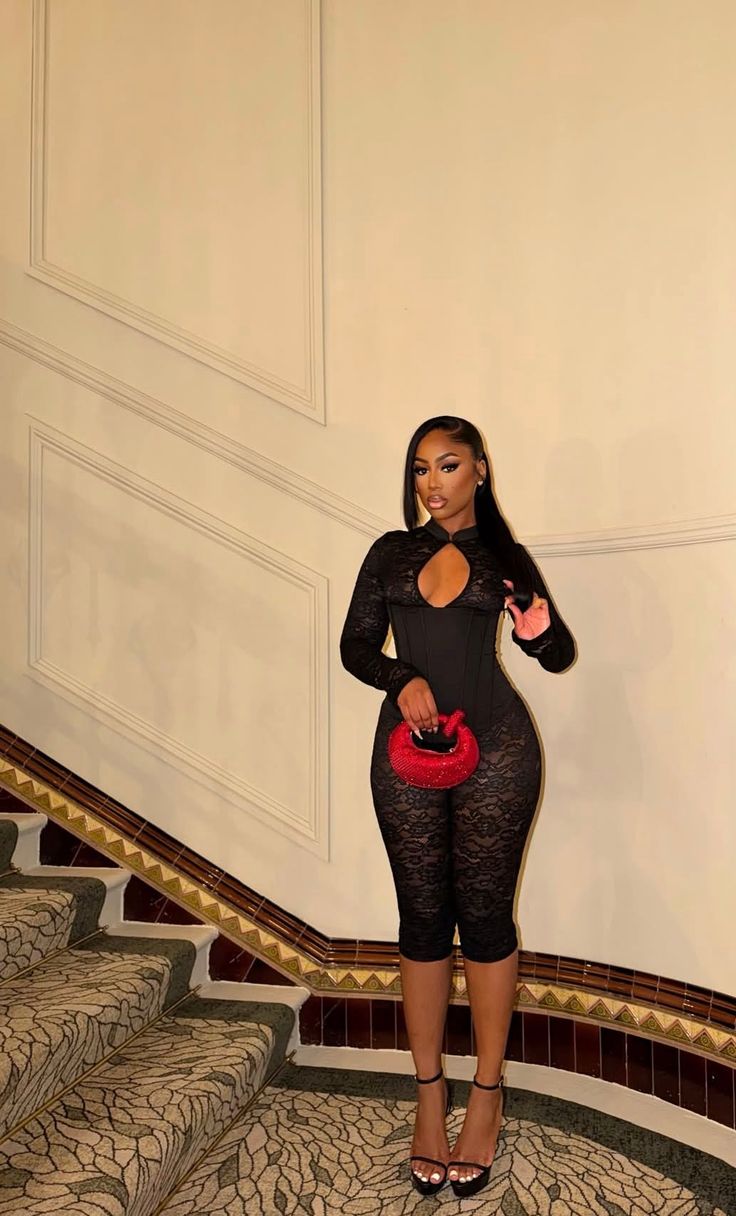 Woman in a black lace jumpsuit and high heels holding a red handbag