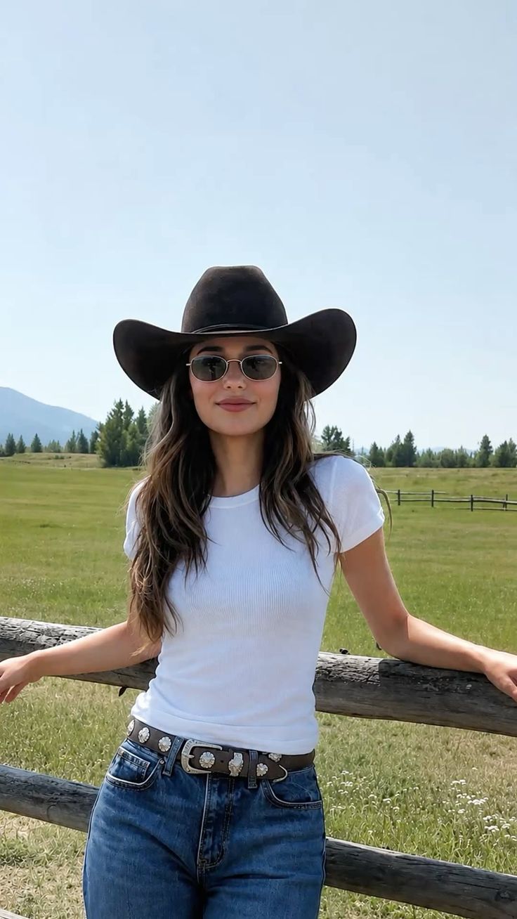 Woman in a cowboy hat and sunglasses leaning on a wooden fence in a green field