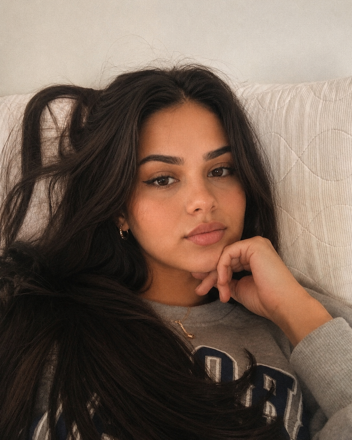 Woman with long dark hair wearing a sweatshirt and jewelry, resting her chin on her hand