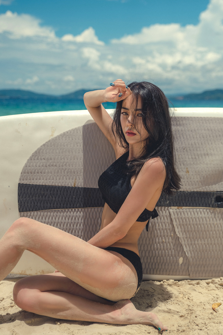 Woman in a black bikini sitting on a sandy beach next to a surfboard