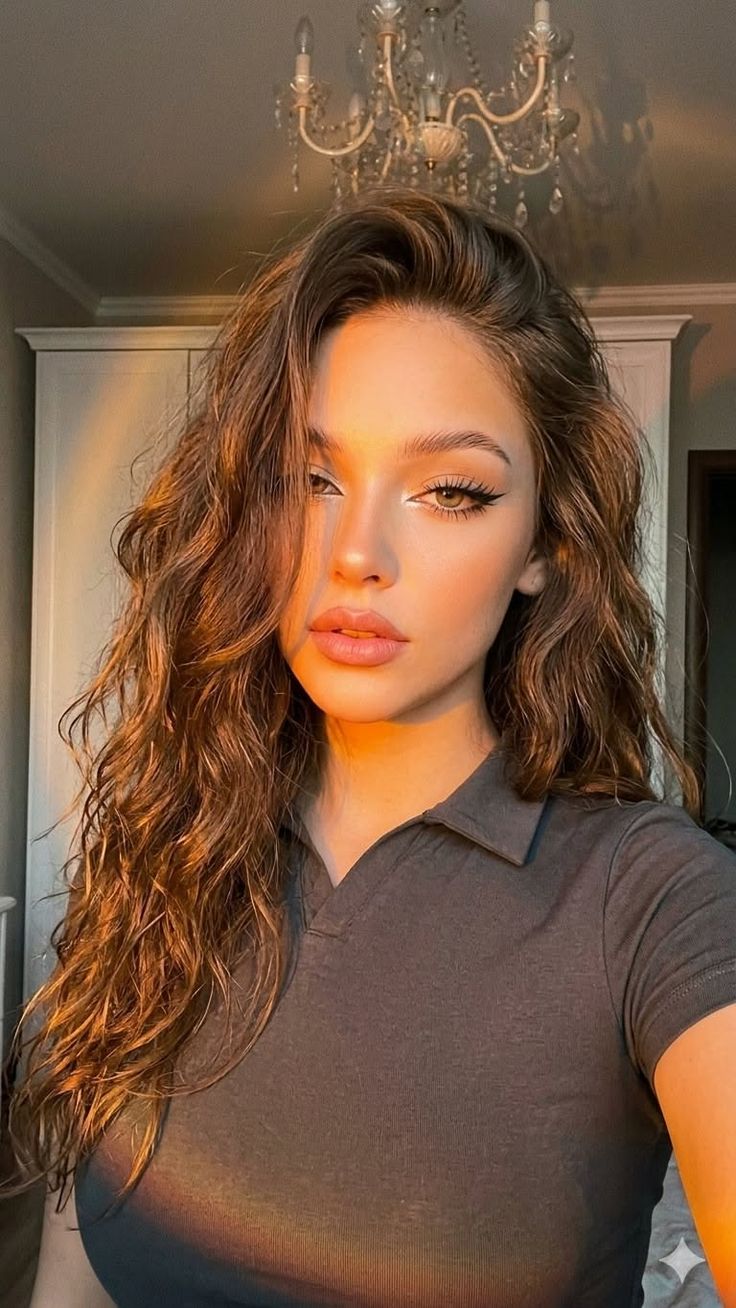 Woman with long wavy hair in a collared shirt, lit by golden hour sunlight indoors