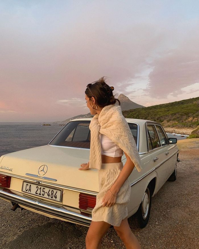 Woman in a knitted set leaning on a vintage car at sunset by the ocean
