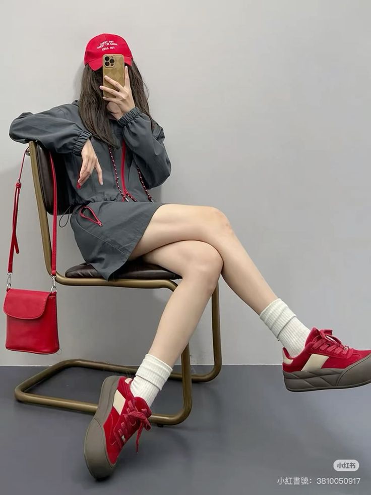 Person in a red cap and gray jacket sitting on a chair, taking a mirror selfie