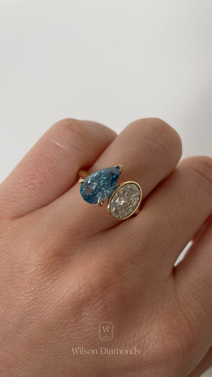 Hand wearing a gold ring with a blue pear-shaped diamond and an oval diamond