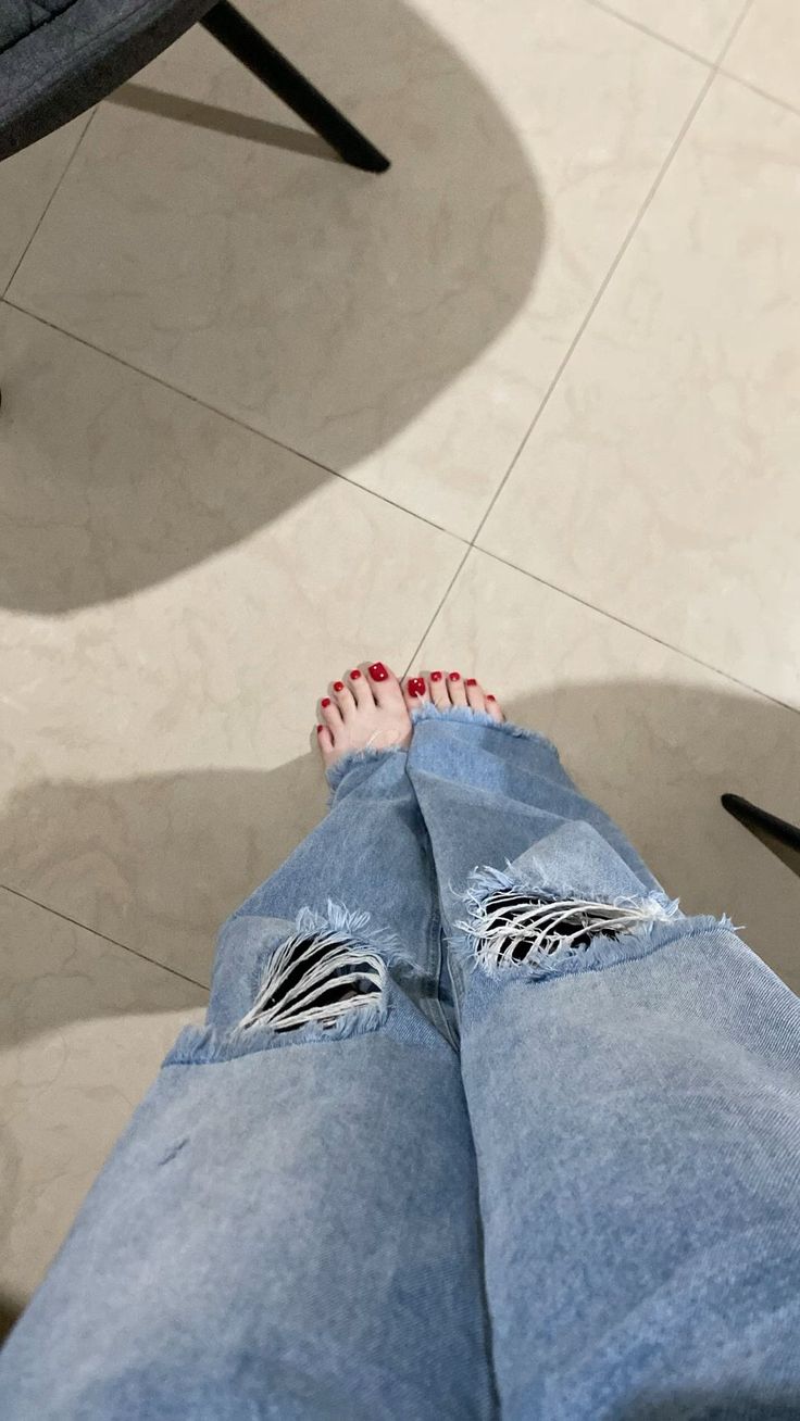 Person wearing ripped jeans with bare feet and red nail polish on a tiled floor